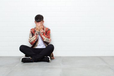 young handsome man feeling sad, frustrated, nervous and depressed, covering face with both hands, crying sitting on cement floor