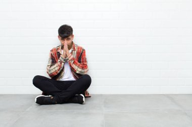 young handsome man feeling worried, hopeful and religious, praying faithfully with palms pressed, begging forgiveness sitting on cement floor