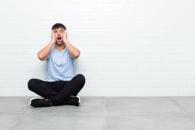 young handsome man looking unpleasantly shocked, scared or worried, mouth wide open and covering both ears with hands sitting on cement floor