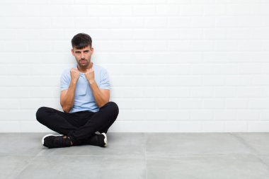 young handsome man looking confident, angry, strong and aggressive, with fists ready to fight in boxing position sitting on cement floor
