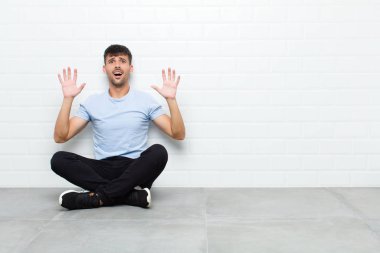 young handsome man feeling stupefied and scared, fearing something frightening, with hands open up front saying stay away sitting on cement floor