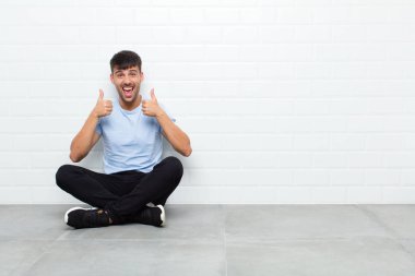 young handsome man smiling broadly looking happy, positive, confident and successful, with both thumbs up sitting on cement floor