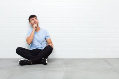 young handsome man thinking, feeling doubtful and confused, with different options, wondering which decision to make sitting on cement floor