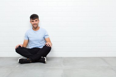 young handsome man smiling cheerfully and casually with a positive, happy, confident and relaxed expression sitting on cement floor