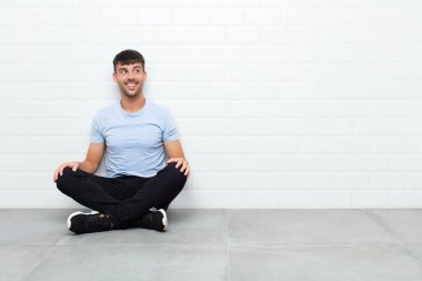 young handsome man feeling shocked, happy, amazed and surprised, looking to the side with open mouth sitting on cement floor