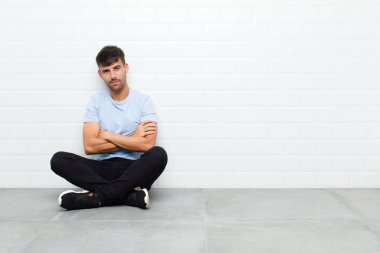 young handsome man feeling displeased and disappointed, looking serious, annoyed and angry with crossed arms sitting on cement floor