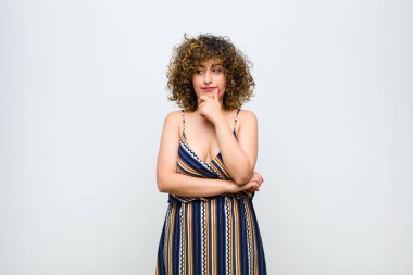 Genç, güzel bir kadın mutlu, kendinden emin bir ifadeyle gülümsüyor. Elleri çenesinde, beyaz duvara bakıyor ve merak ediyor.