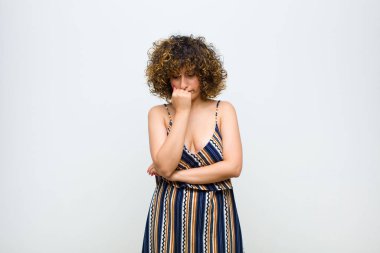 Genç ve güzel bir kadın ciddi, düşünceli ve endişeli, elleri beyaz duvara yaslanmış bir şekilde yan bakıyor.