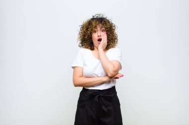 Genç ve güzel bir kadın şok ve inançsızlık içinde, elleri yanak ve kolları çaprazlanmış, sersemlemiş ve beyaz duvara karşı hayrete düşmüş.