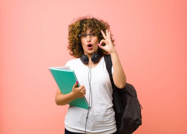 Genç, güzel bir bayan öğrenci kendini mutlu, şaşkın, tatmin olmuş ve şaşırmış hissediyor, iyi olduğunu gösteriyor ve işaret ediyor, pembe duvara gülümsüyor.
