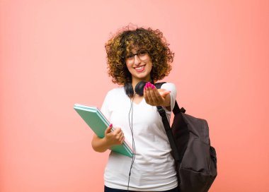 Genç ve güzel bir kız öğrenci mutlu bir şekilde gülümsüyor. Dost canlısı, kendinden emin, pozitif bakışlı, pembe duvara karşı bir nesne ya da konsept sunuyor ve gösteriyor.