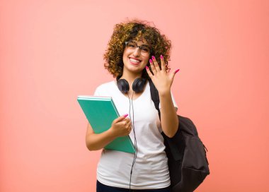 Genç ve güzel bir bayan öğrenci gülümsüyor ve arkadaş canlısı görünüyor, 5 ya da 5 numarayı gösteriyor, elini ileri uzatmış, pembe duvara doğru sayıyor.