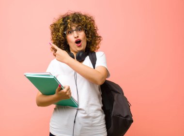 Genç ve güzel bayan heyecanlı ve şaşırmış görünüyor. Pembe duvara uzayı kopyalamak için yan ve yukarı işaret ediyor.