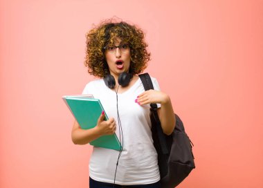 Genç ve güzel bir kız öğrenci ağzı açık, şaşkın ve şaşkın görünüyor, pembe duvara karşı kendini işaret ederek.