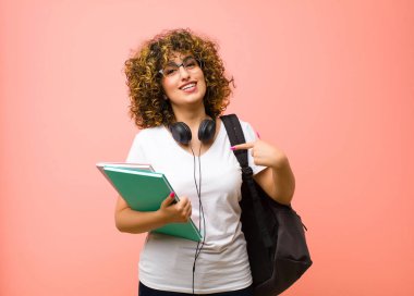 Genç ve güzel bir bayan gururlu, kendinden emin ve mutlu görünüyor, gülümsüyor ve kendini işaret ediyor ya da pembe duvara karşı bir numara işareti yapıyor.