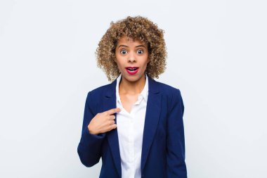 Afro-Amerikan görünümlü genç bir kadın ağzı açık, şok olmuş ve şaşırmış görünüyor, düz duvara karşı kendini işaret ederek