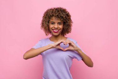 Genç Afrikalı Amerikalı kadın gülümsüyor ve mutlu, sevimli, romantik ve aşık, iki elini de pembe duvara dayayıp kalp şeklinde yapıyor.