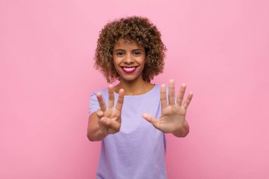 Genç Afrikalı Amerikalı kadın gülümsüyor ve arkadaş canlısı görünüyor, 8 ya da 8 numarayı gösteriyor, elleri önde, pembe duvara doğru sayıyor.