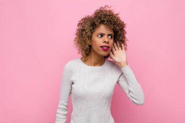 Genç Afrikalı Amerikalı kadın ciddi ve meraklı görünüyor, dinliyor, gizli bir konuşma veya dedikodu duymaya çalışıyor, pembe duvara karşı kulak misafiri oluyor.