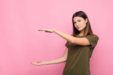 Genç, güzel bir kadın gülümsüyor, mutlu, pozitif ve tatmin olmuş hissediyor, fotokopi odasında pembe duvara karşı nesne ya da konsept tutuyor ya da gösteriyor