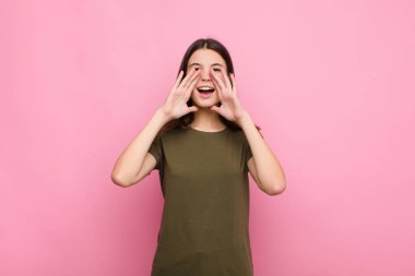 Genç, güzel bir kadın mutlu, heyecanlı ve pozitif hissediyor. Elleri ağızda, pembe duvara doğru bağırıyor.