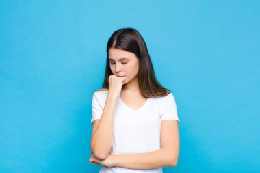 Genç ve güzel bir kadın ciddi, düşünceli ve endişeli, elleri mavi duvara yaslanmış bir şekilde yan bakıyor.