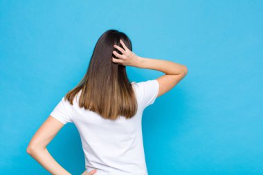 Genç güzel bir kadın düşünüyor ya da şüphe ediyor, kafasını kaşıyor, şaşkın ve şaşkın hissediyor, mavi duvarın arkasında ya da arkasında.