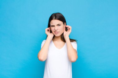 Genç, güzel bir kadın sinirli, stresli ve sinirli görünüyor, her iki kulağı da sağır edici bir gürültü, ses veya yüksek sesle kaplayan mavi duvara karşı müzik