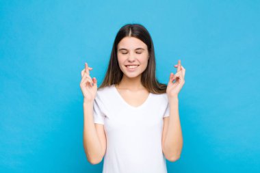 Genç, güzel bir kadın gülümsüyor ve endişeyle iki parmağını çapraz yapıyor. Endişeli hissediyor ve mavi duvara karşı iyi şanslar diliyor veya umut ediyor.