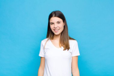 Genç ve güzel bir kadın neşeli bir şekilde gülümsüyor, pozitif, mutlu, kendinden emin ve gevşek bir ifadeyle mavi duvara karşı.