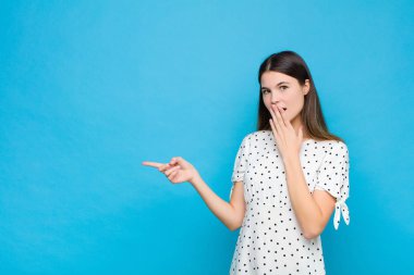 Genç, güzel bir kadın kendini mutlu, şaşkın ve şaşırmış hissediyor. Ağzını eliyle kapatıyor ve mavi duvara karşı yanal fotokopi alanını işaret ediyor.