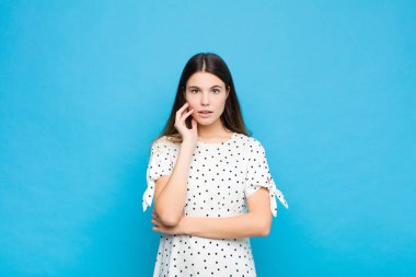 Genç ve güzel bir kadın şok ve inançsızlık içinde, elleri yanağında ve kolları çaprazlanmış, sersemlemiş ve mavi duvara karşı şaşkınlık içinde.