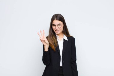 Genç iş kadını gülümsüyor ve arkadaş canlısı görünüyor. Üç ya da üç numarayı gösteriyor. Elleri önde, beyaz duvara doğru sayıyor.