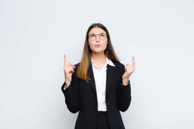 Genç iş kadını şaşırmış, şaşırmış ve ağzı açık görünüyor, iki eliyle yukarıya doğru uzayı beyaz duvara kopyalamak için işaret ediyor.