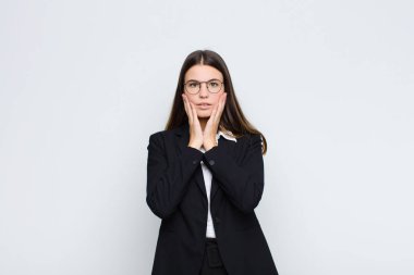 Genç iş kadını şok olmuş ve korkmuş hissediyor. Ağzı açık ve elleri beyaz duvarda.