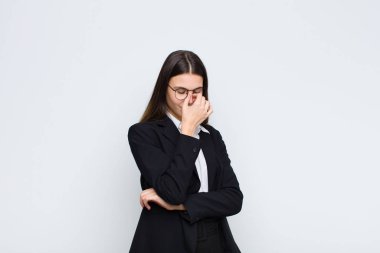 Genç iş kadını kendini stresli, mutsuz ve hayal kırıklığına uğramış hissediyor, alnına dokunuyor ve beyaz duvara karşı şiddetli baş ağrısı çekiyor.