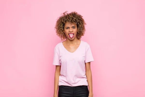 若いアフリカ系アメリカ人の女性はうんざりしていらいらして舌を出しピンクの壁に対して何か厄介で幸運を嫌い — ストック写真