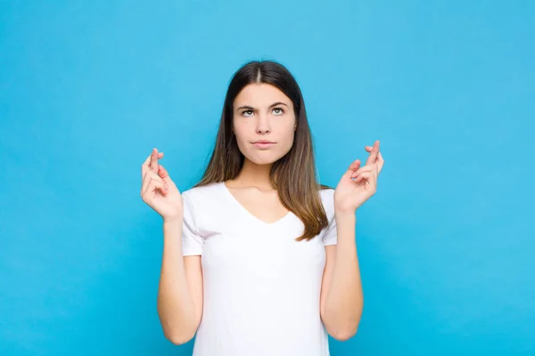 young pretty woman crossing fingers anxiously and hoping for good luck with a worried look against blue wall