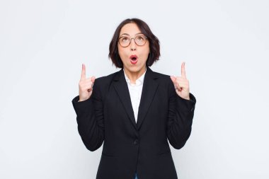 Genç iş kadını huşu ve açık ağızla beyaz duvara karşı şaşkın ve şaşkın bir bakışla yukarıya bakıyor.