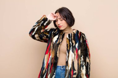 Genç ve güzel bir kadın stresli görünüyor, yorgun ve hayal kırıklığına uğramış, alnından ter damlıyor, bej rengi duvara karşı umutsuz ve bitkin hissediyor.