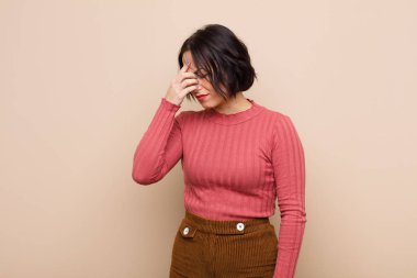 Genç, güzel bir kadın stresli, mutsuz ve hayal kırıklığına uğramış, alnına dokunmuş ve bej rengi duvara karşı şiddetli baş ağrısı çeken.