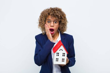 Genç Afrikalı Amerikalı kadın şok olmuş ve korkmuş hissediyor. Ağzı açık ve elleri ev modeliyle yanakları üzerinde korkmuş görünüyor.