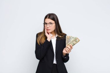 Genç ve güzel bir kadın şok olmuş ve korkmuş hissediyor. Ağzı açık ve elleri banknotlarla yanaklarında korkmuş görünüyor.