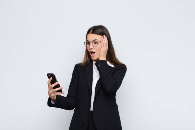 Genç, güzel bir kadın mutlu, heyecanlı ve şaşırmış hissediyor, iki eli yüzü üzerinde bir cep telefonuyla yan tarafa bakıyor.