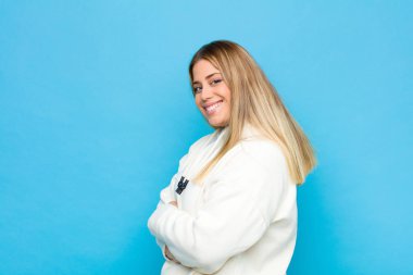 Genç sarışın kadın kameraya çapraz kollarıyla gülümsüyor ve mutlu, kendinden emin, tatmin olmuş bir yüz ifadesi düz duvara karşı yanal bir bakış.