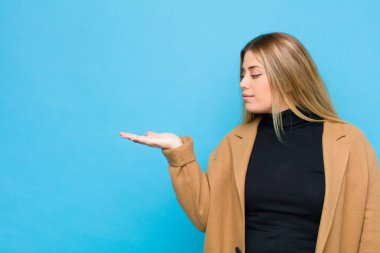 Genç sarışın kadın kendini mutlu hissediyor ve gelişigüzel gülümsüyor. Bir objeye ya da duvara yaslanmış bir konsepte bakıyor.