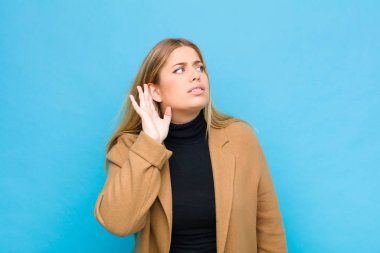 Genç sarışın kadın ciddi ve meraklı görünüyor, dinliyor, gizli bir konuşma ya da dedikodu duymaya çalışıyor, düz duvara kulak misafiri oluyor.