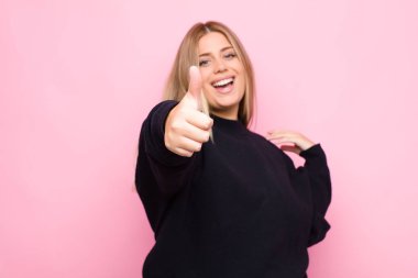 Genç sarışın kadın gururlu, kaygısız, kendinden emin ve mutlu, parmakları düz duvara değmiş bir şekilde gülümsüyor.