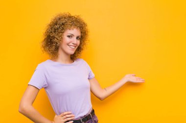 Genç afro kadın kendini mutlu ve neşeli hissediyor, gülümsüyor ve seni selamlıyor, turuncu duvara karşı dostça bir jestle içeri davet ediyor.