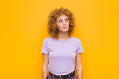 Endişeli, kafası karışık, budala bir ifadeyle afro genç bir kadın, kopya alanı arıyor, turuncu duvara karşı şüpheyle bakıyor.